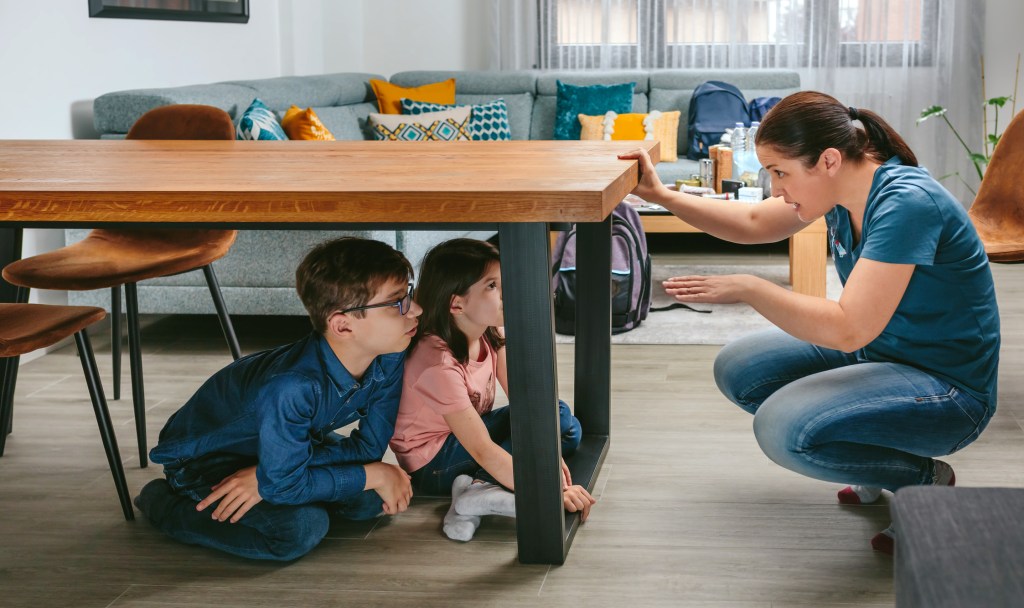 Mother explaining to her children how to protect themselves in an earthquake by getting under the table while preparing emergency backpacks