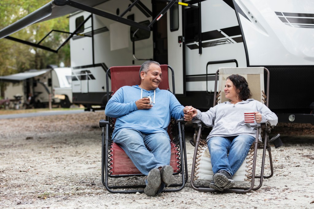 A couple sitting together in chairs by a camper trailer in an RV park, drinking coffee.