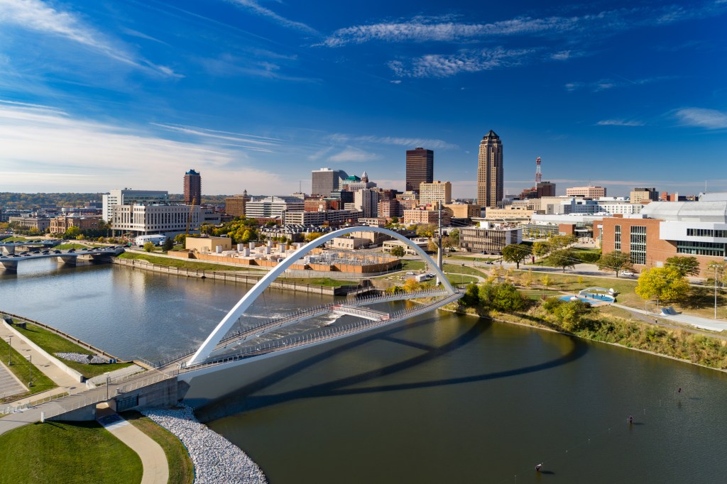 Aerial of the Des Moines Riverwalk bridge and the Des Moines River, with the Downtown Des Moines skyline in the background.