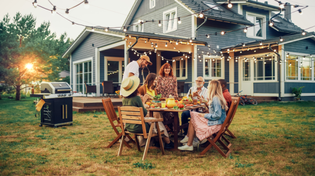 A family gathers around a wooden table for an outdoor dinner on a lawn at dusk under string lights, with a house in the background.